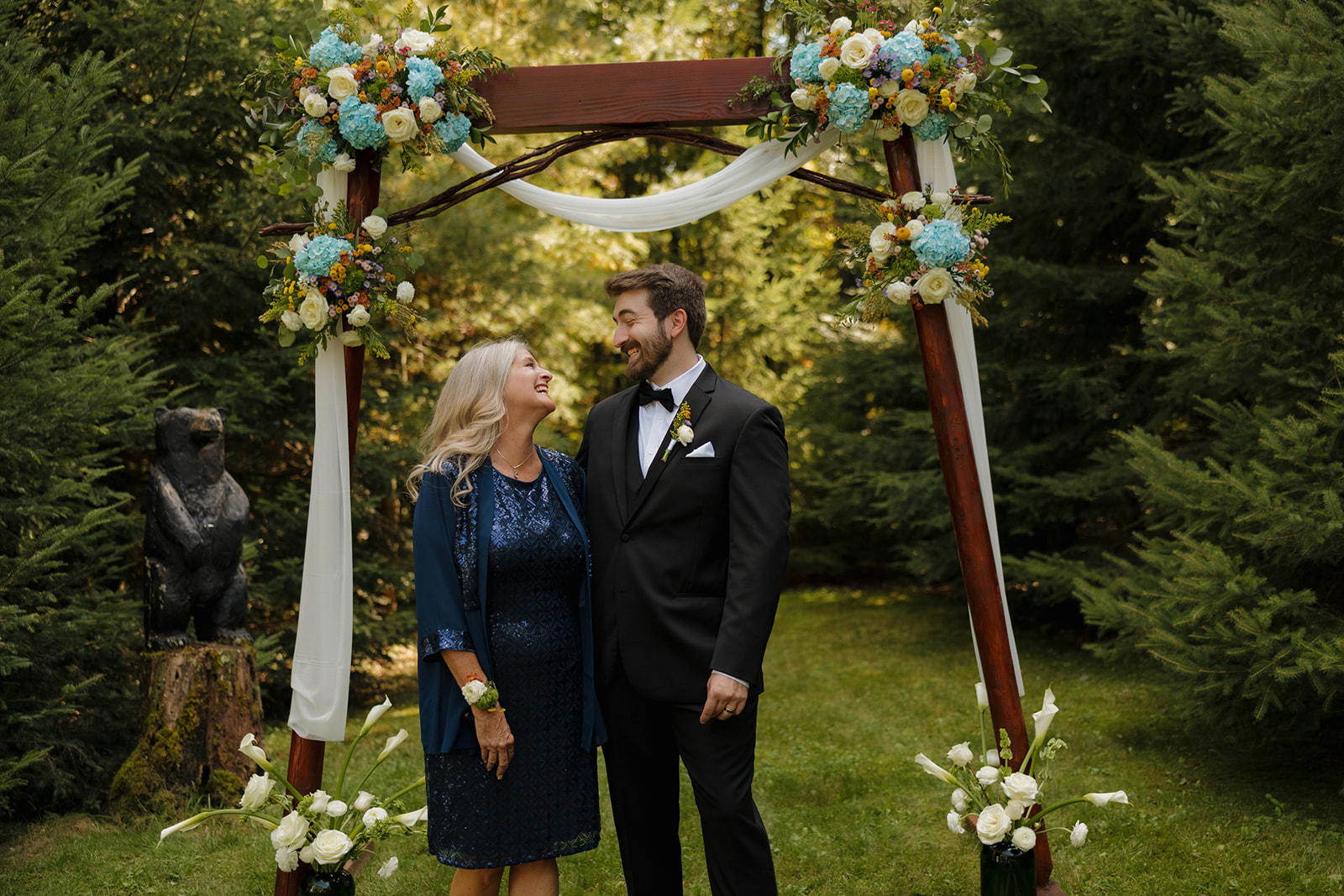 Groom smiling with his mother beneath the wooden arch decorated with flowers.