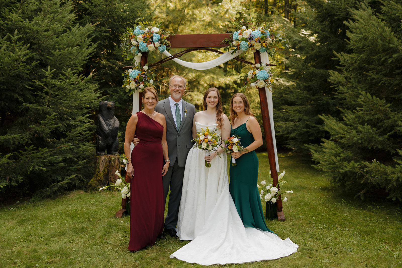 Bride posing with her parents and sister beneath a floral ceremony arch at a barn wedding.