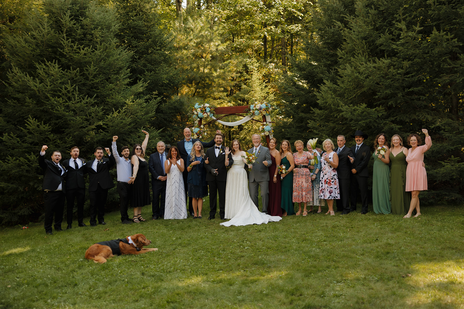 Large group family photo with the bride and groom under the floral arch, guests cheering around them.