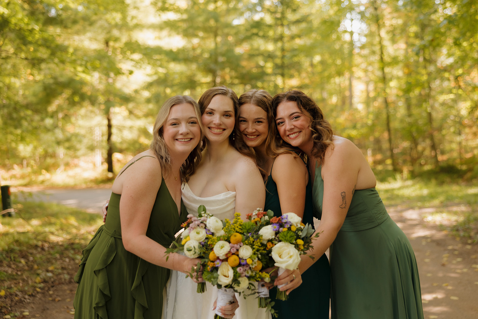 Bride smiling with her bridesmaids in green dresses, holding a colorful bouquet at a barn wedding.