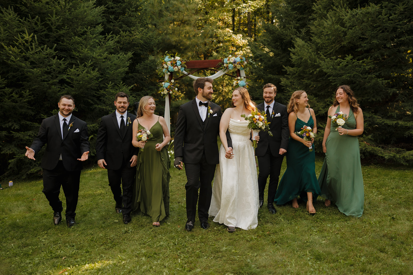 Wedding party walking together on the grass, bridesmaids in green dresses and groomsmen in black suits at a farm wedding.