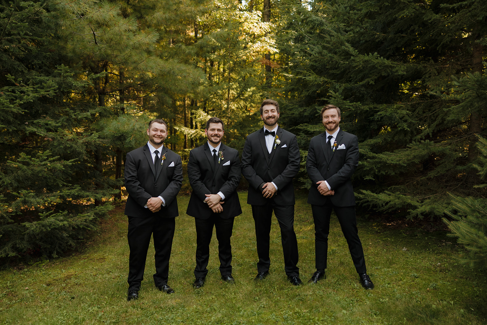 Groom standing with his groomsmen in black tuxedos surrounded by tall pines.
