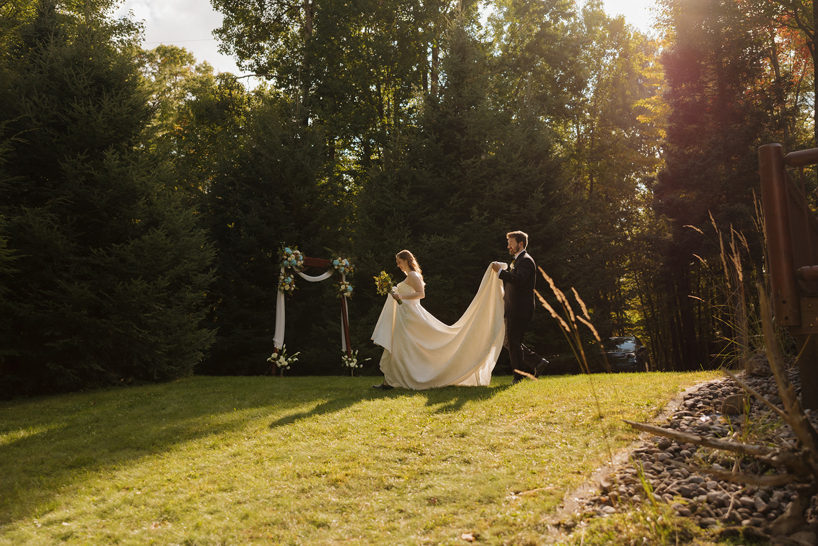 Bride and groom walking across a sunny lawn, groom carrying the bride’s train as the ceremony arch waits behind them.