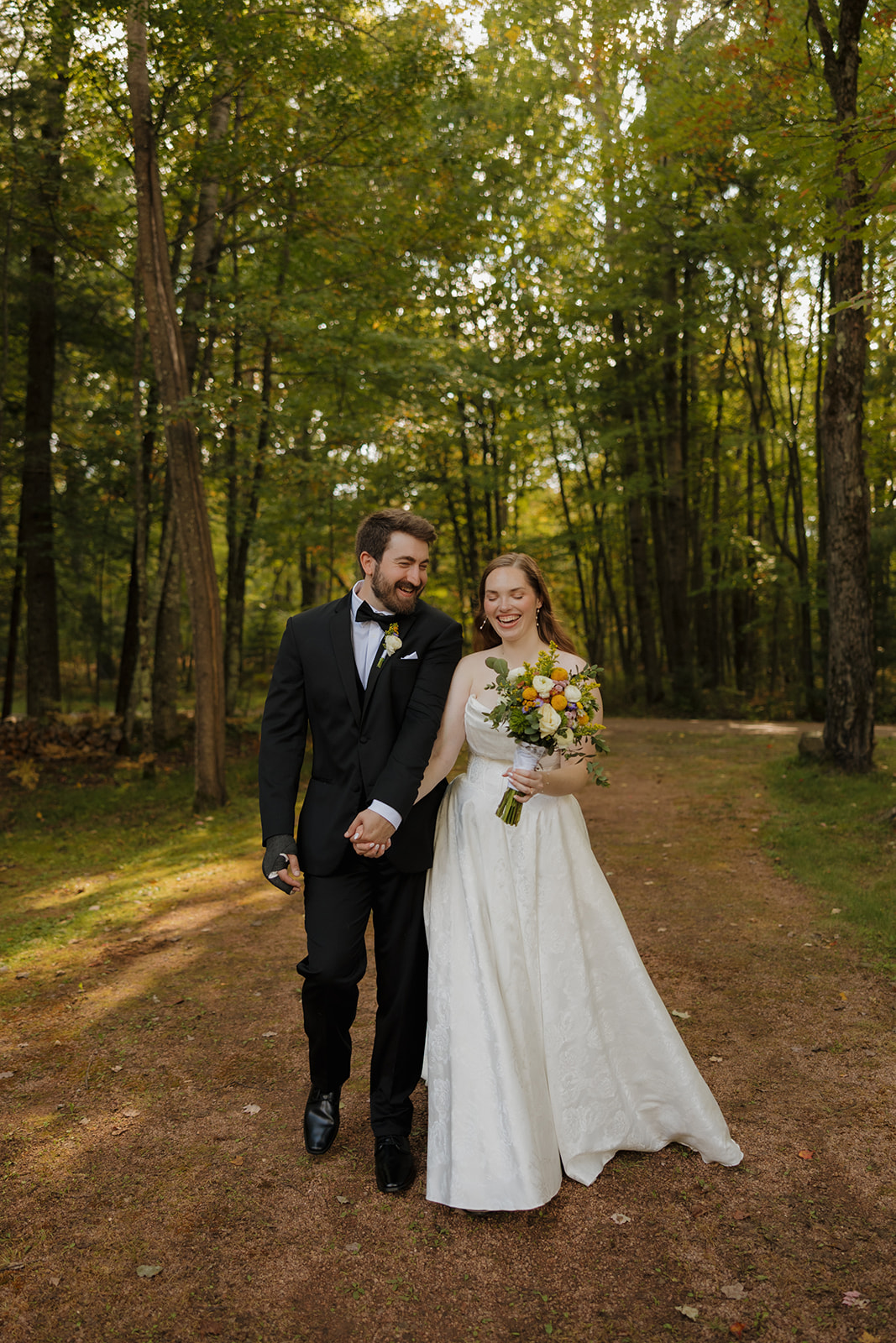 Couple walking hand in hand down a wooded path, bride holding a bright bouquet.