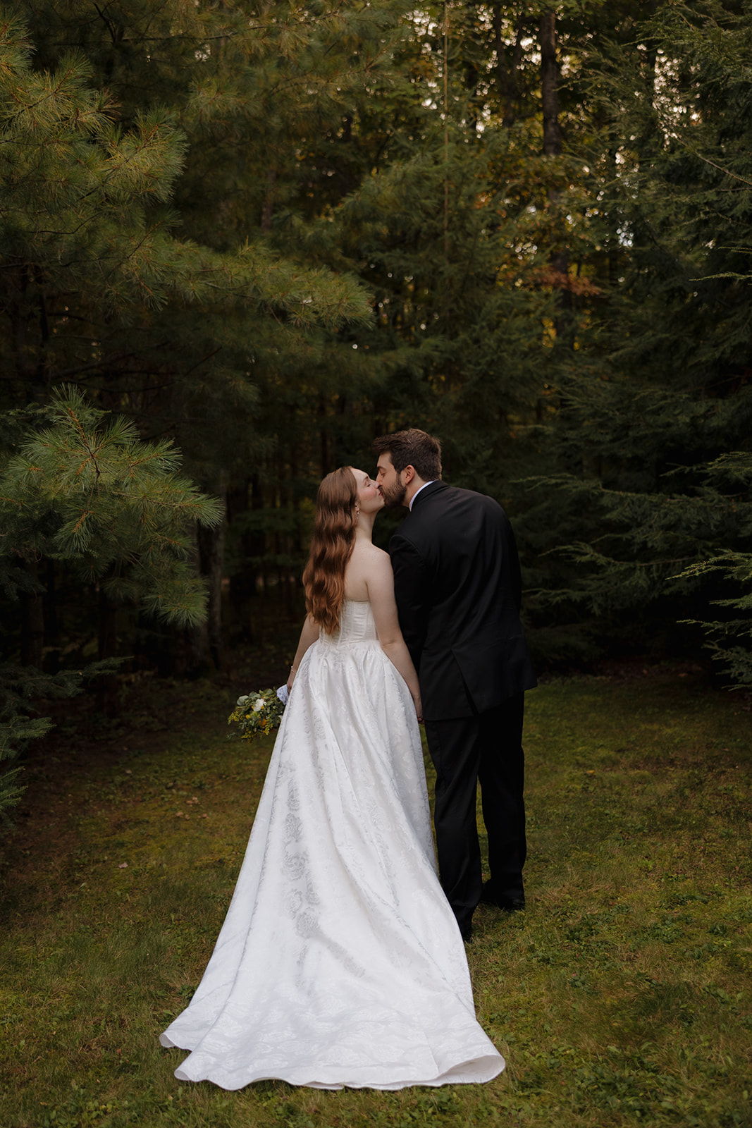 Newlyweds kiss among tall pine trees, the bride’s long gown flowing behind her.