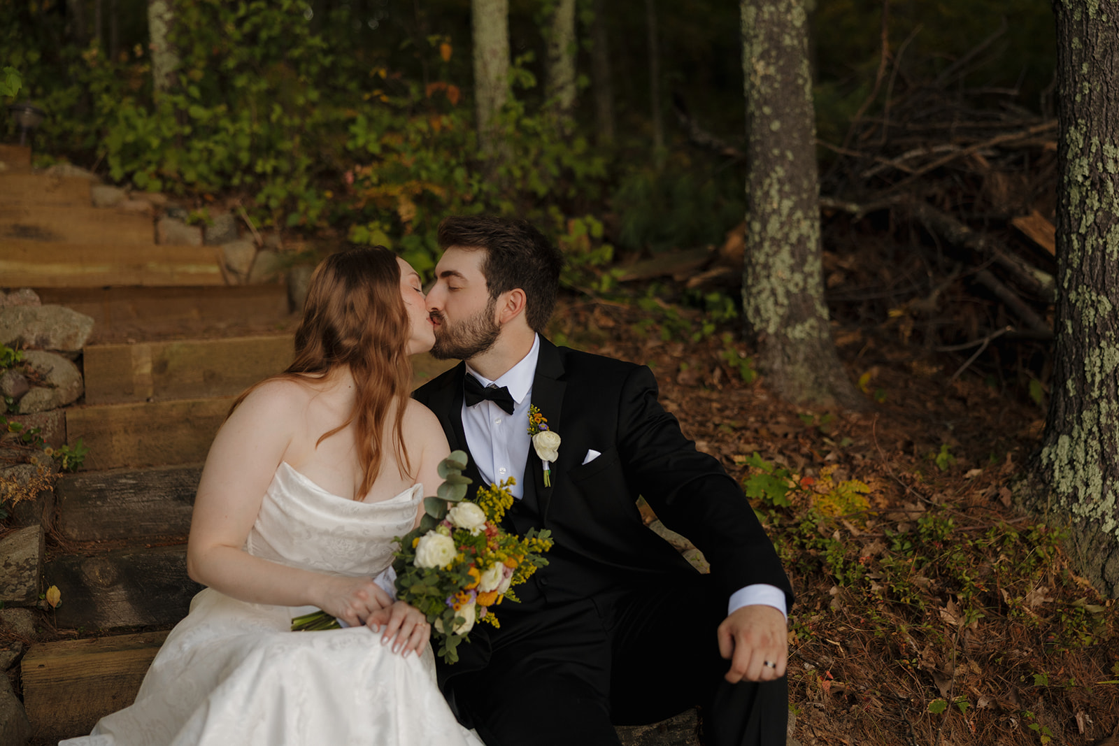 Bride and groom share a kiss on rustic stone steps surrounded by trees at their barn wedding.