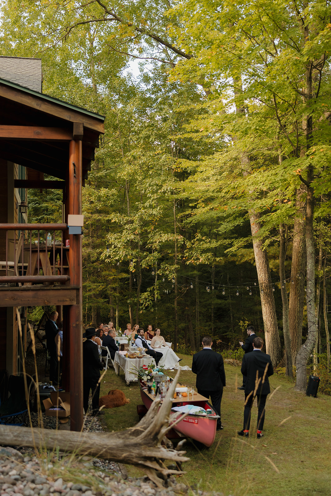 Outdoor wedding reception beside a rustic cabin, with long tables and string lights among the trees.