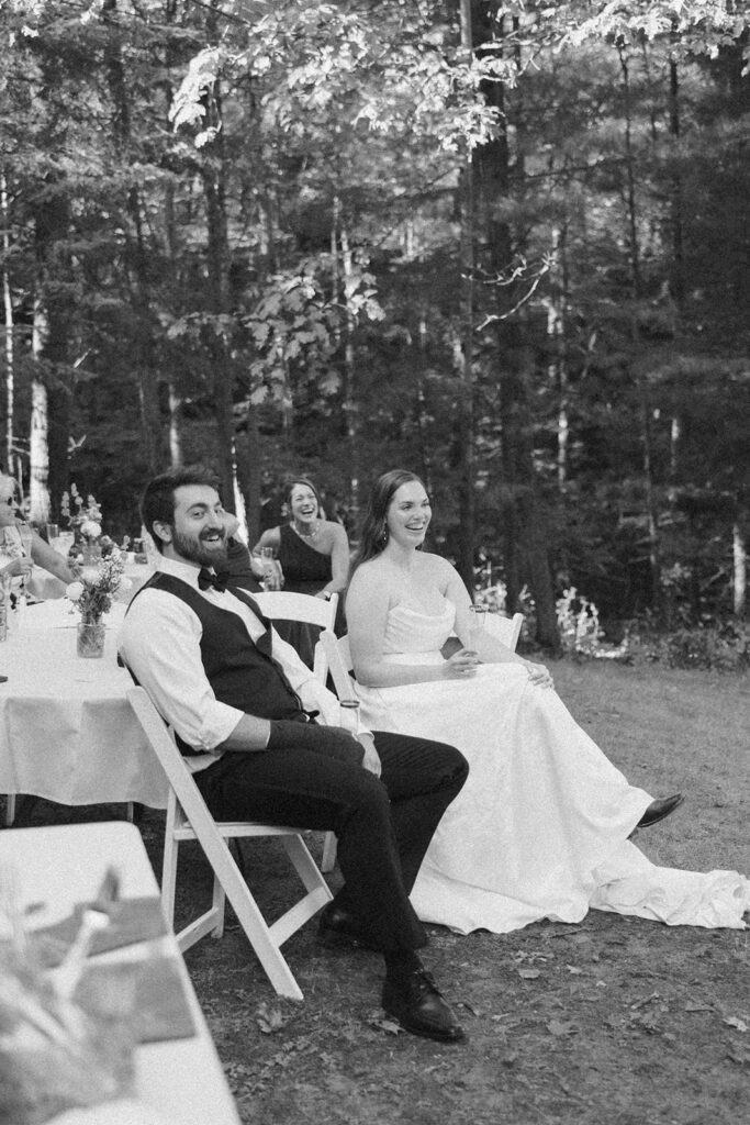 Black and white candid of bride and groom smiling during reception speeches outdoors.