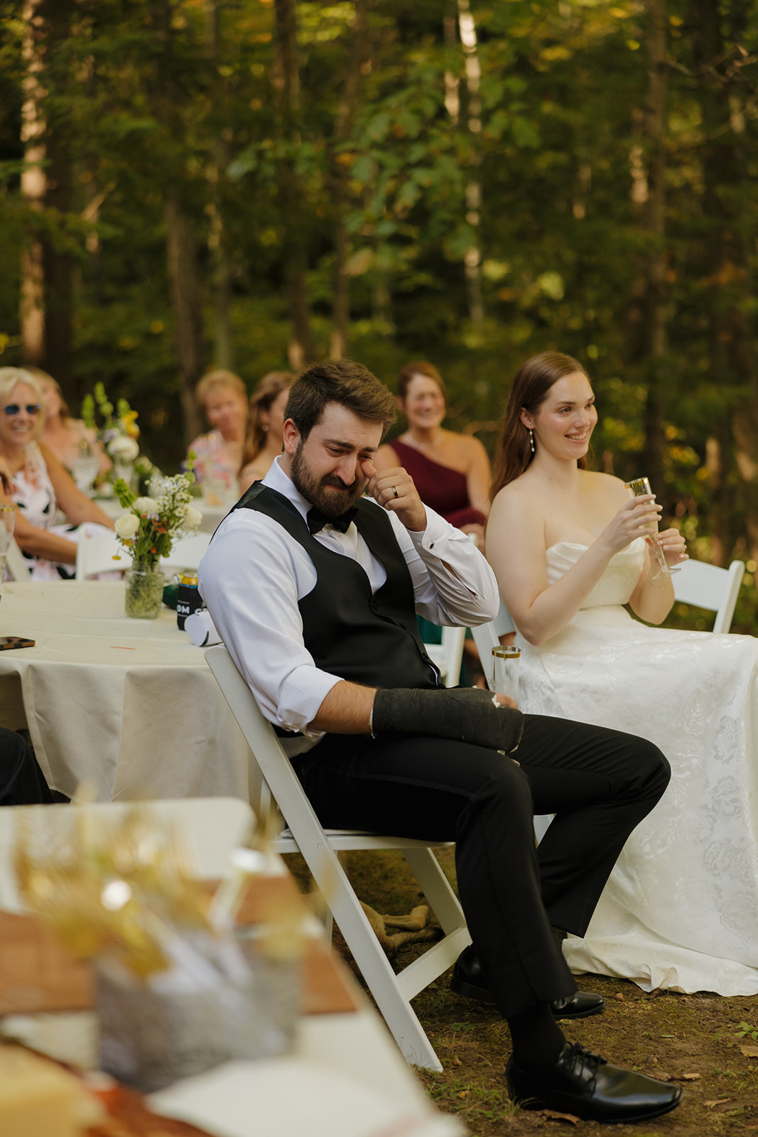 Groom wiping away tears while seated during reception speeches at a farm wedding, bride smiling beside him.