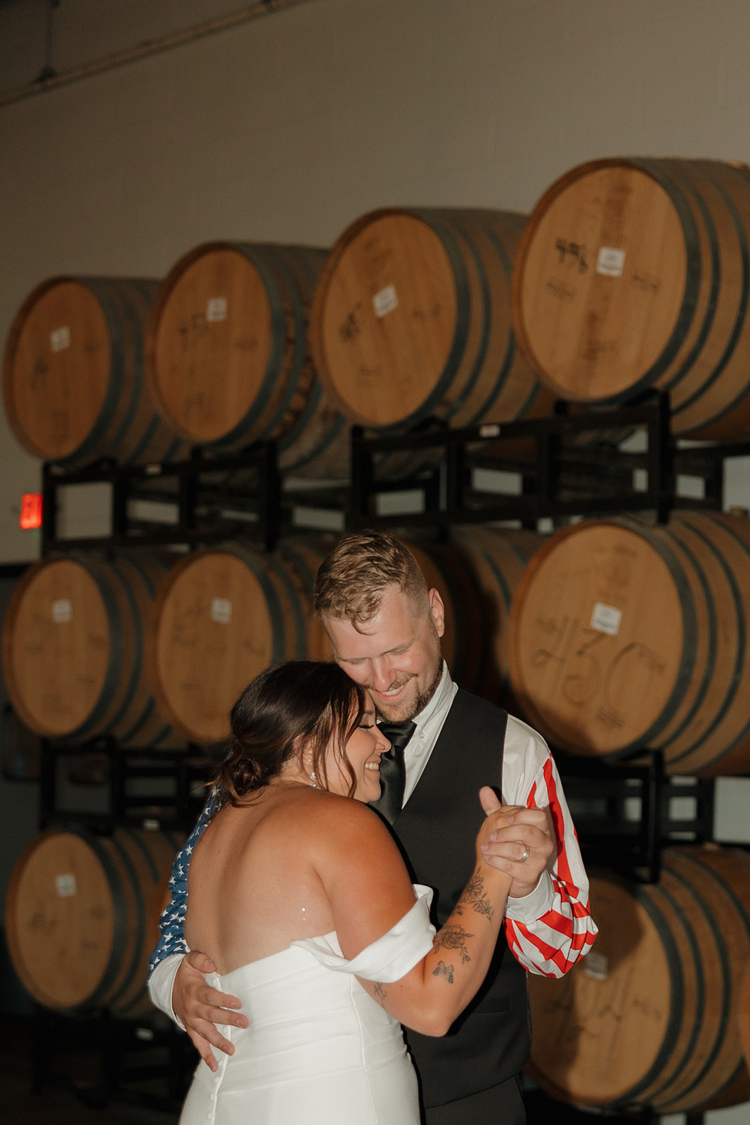 Bride rests her head on the groom’s shoulder as they sway together in front of stacked barrels, soaking in a tender moment during their Wisconsin brewery wedding reception.