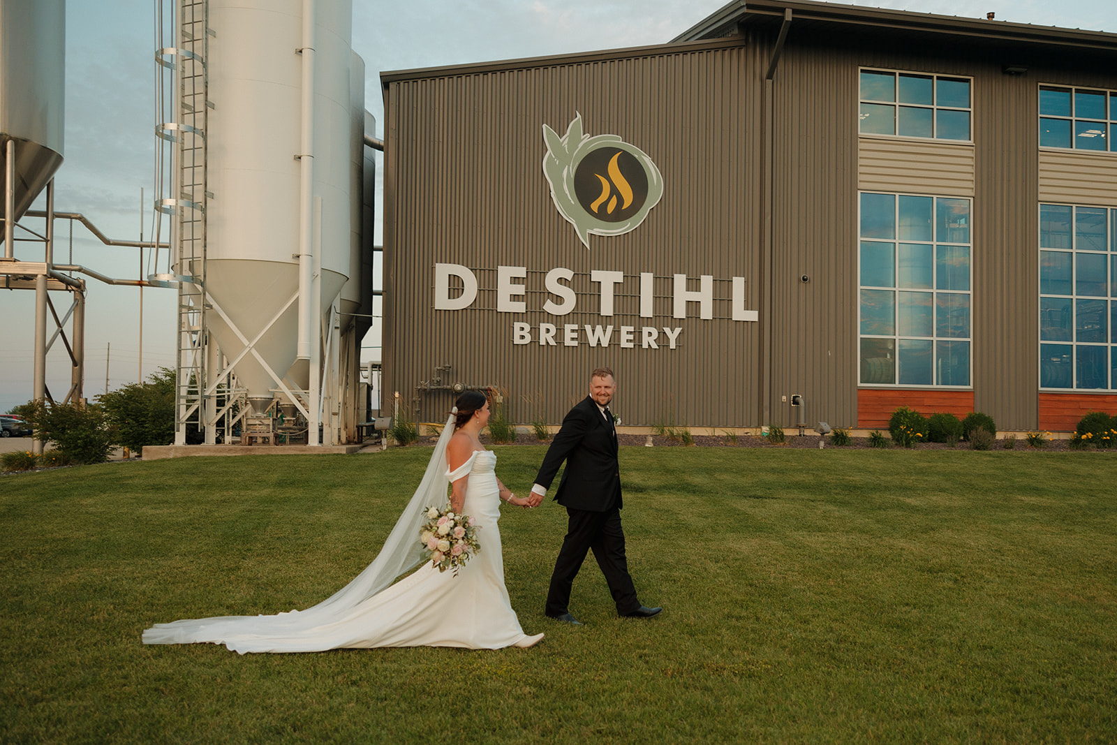 Bride and groom walk hand in hand across the lawn with the iconic DESTIHL Brewery building as their backdrop—quintessential Wisconsin brewery wedding photo.