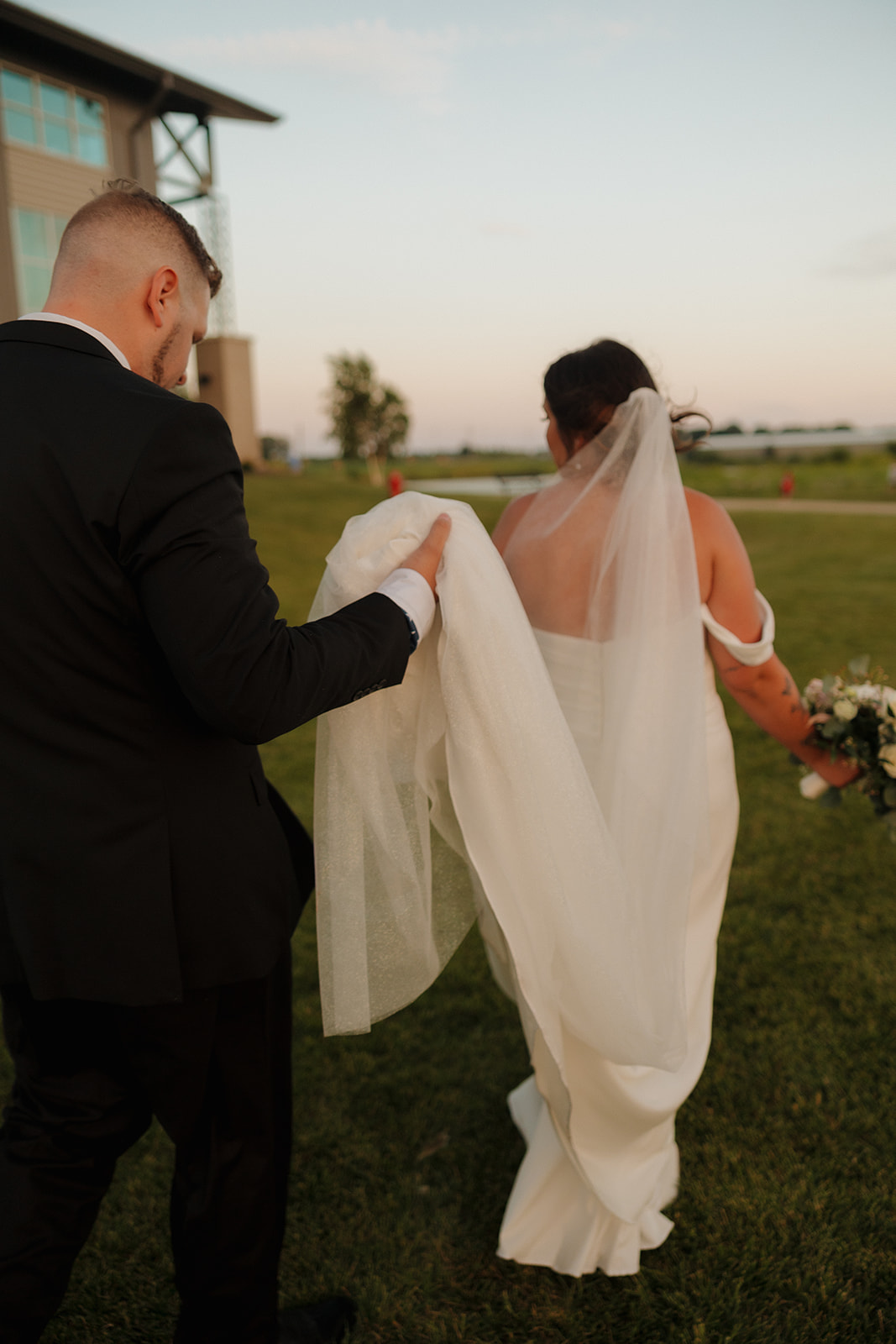 Groom helps carry the bride’s veil as they walk together into golden hour glow outside their Wisconsin brewery reception.