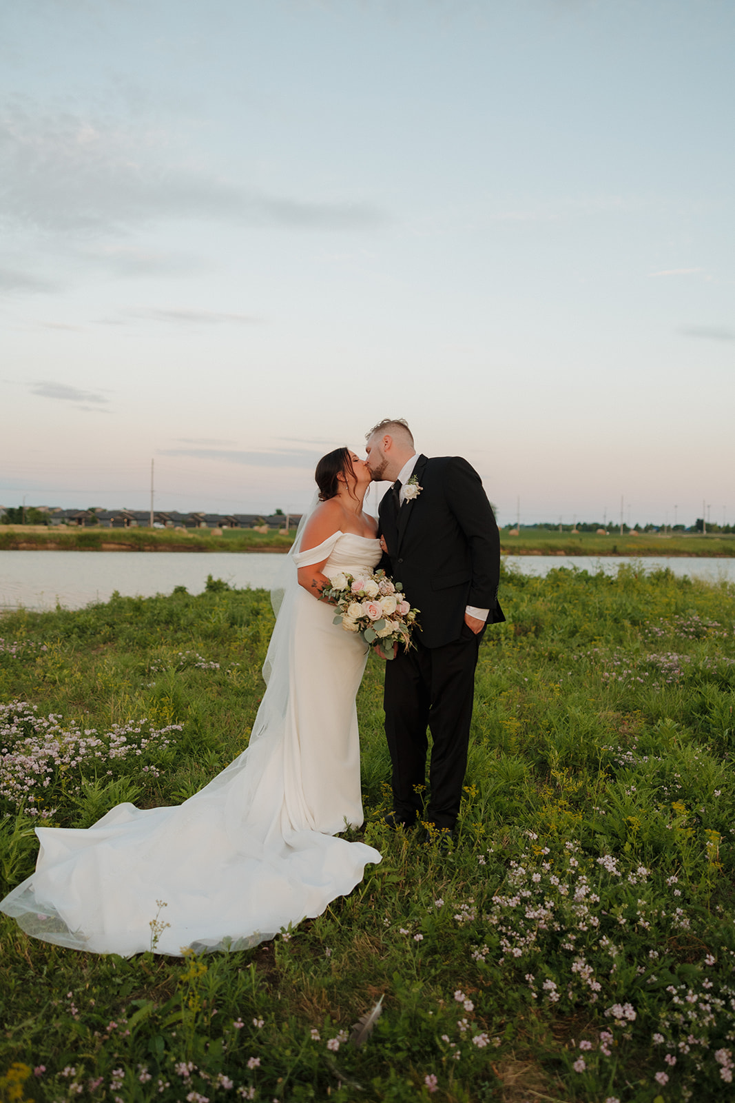 Bride and groom share a kiss in a field of wildflowers near the water at sunset, just steps from their Wisconsin brewery venue.