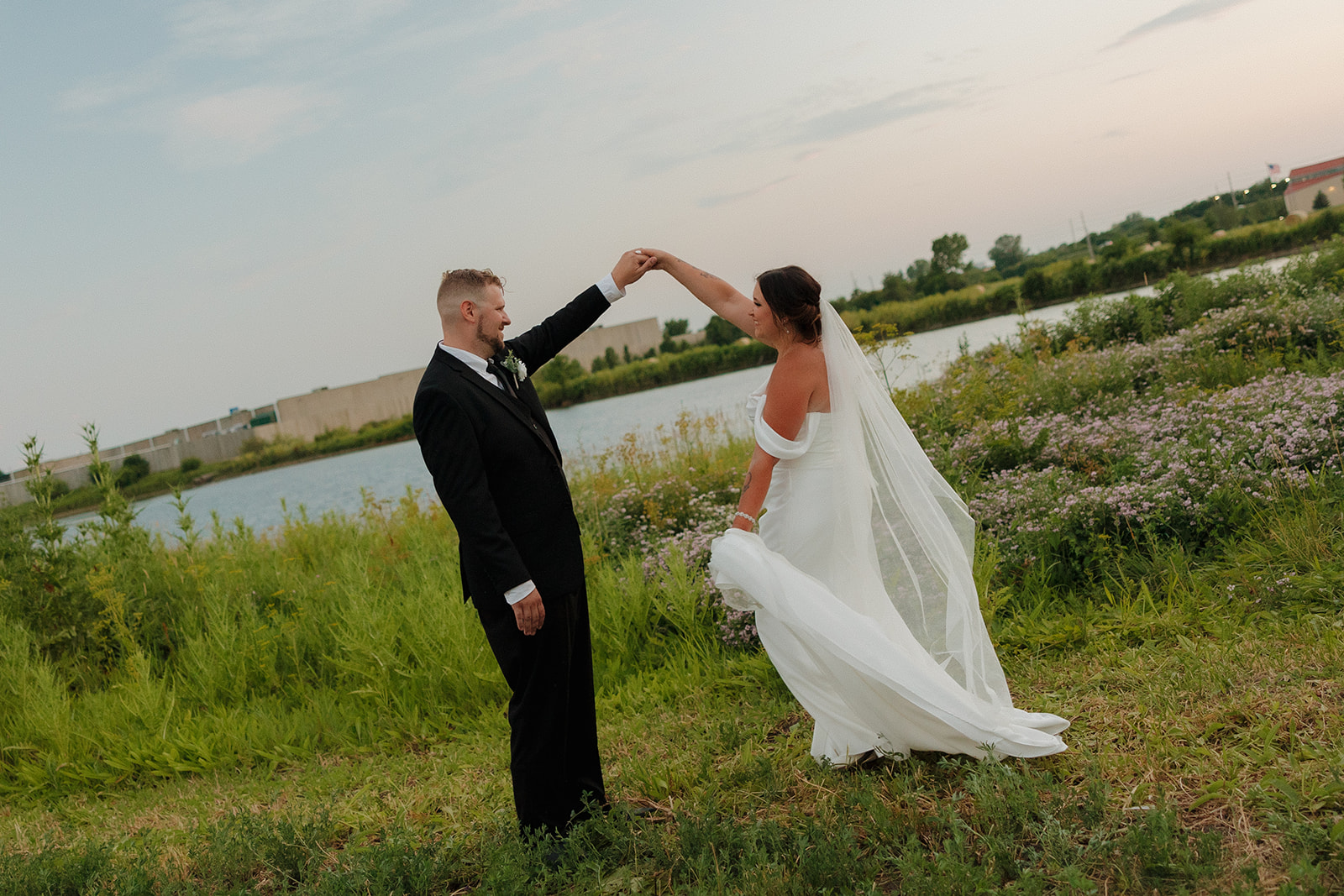 Groom twirls his bride in her off-the-shoulder gown during golden hour by the water near their Wisconsin brewery wedding venue.
