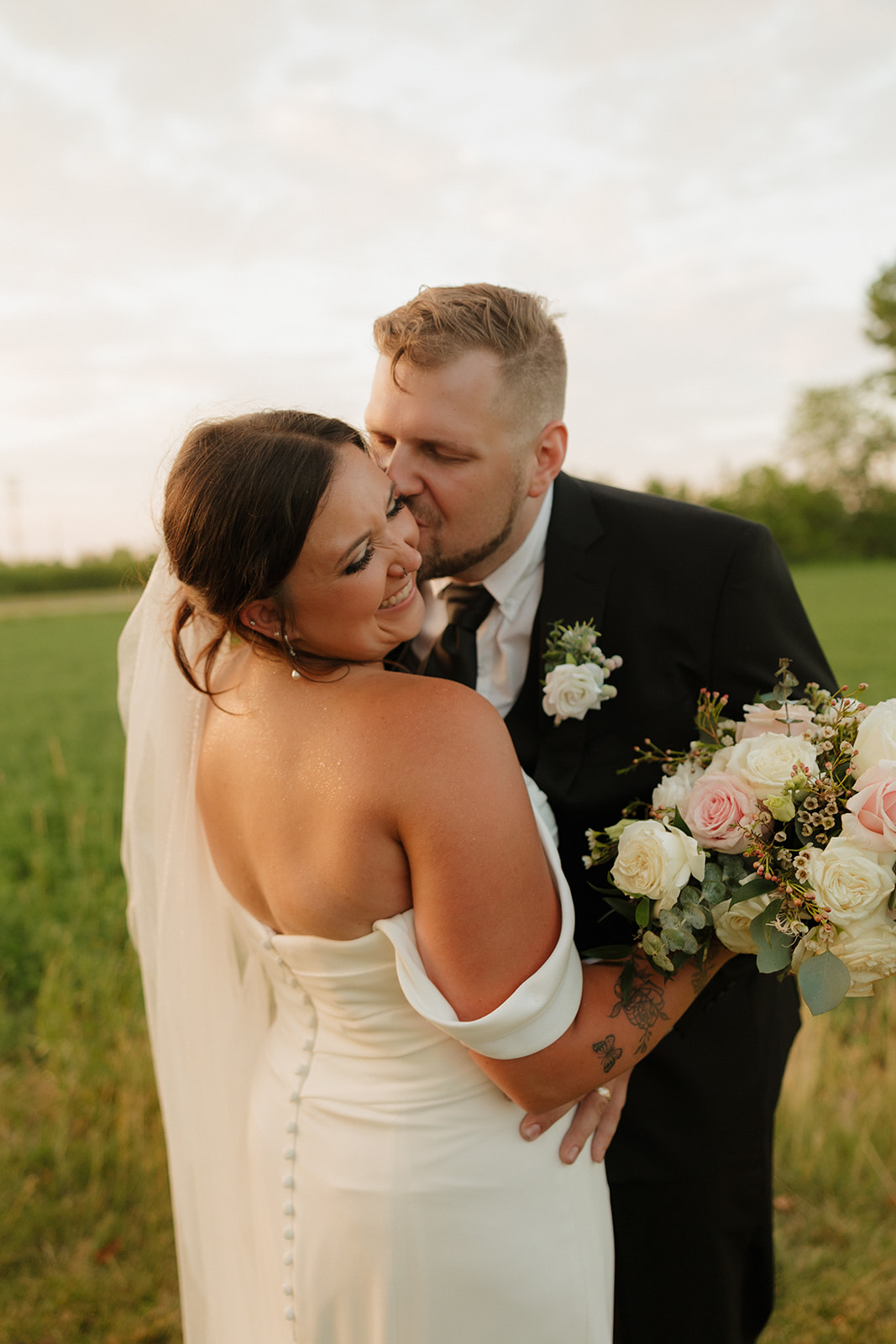 Groom kisses the bride’s cheek as she beams, holding a romantic bouquet during golden hour at their Wisconsin brewery wedding.