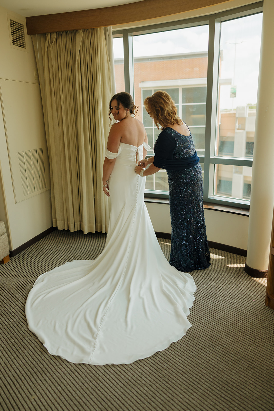 Bride smiles over her shoulder while her mother helps zip her classic white gown, getting ready at a Wisconsin brewery wedding.