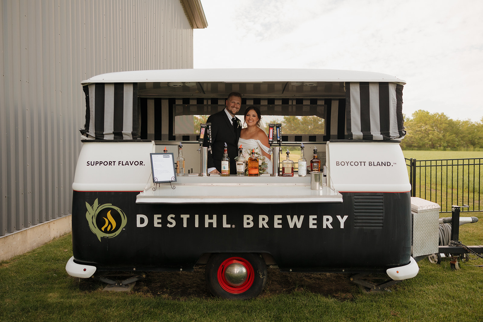 Bride and groom stand behind a custom mobile bar labeled "Destihl Brewery," ready to pour drinks at their Wisconsin brewery reception.
