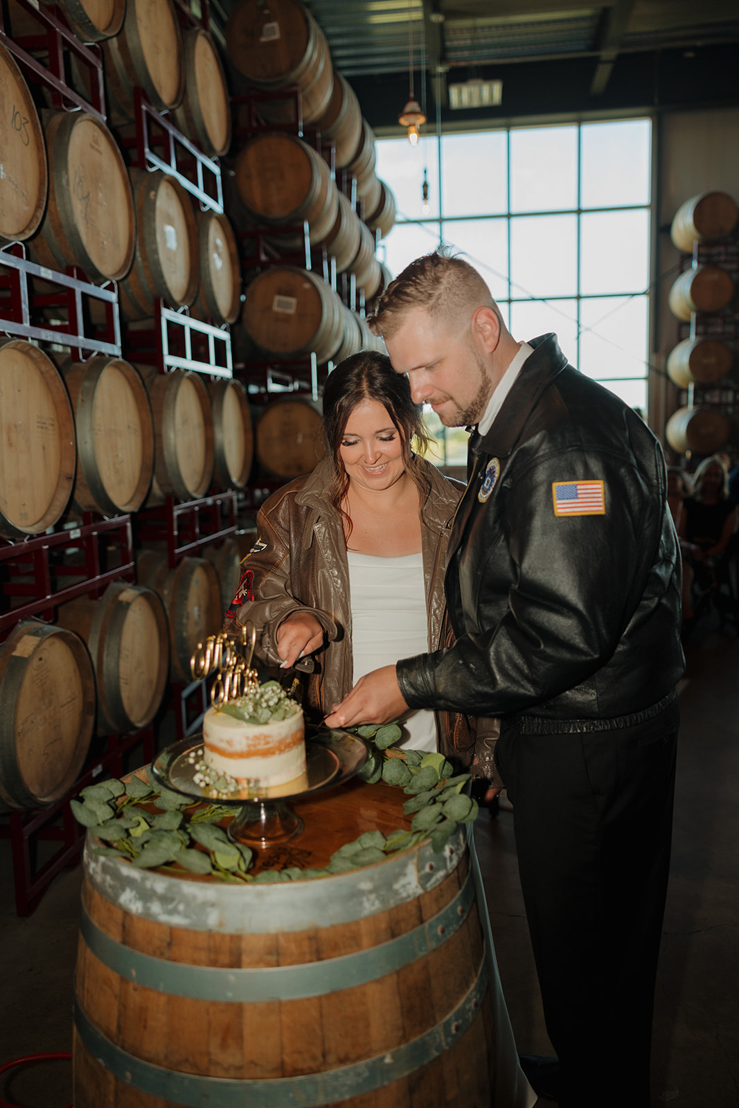 Bride and groom in matching leather jackets cut their wedding cake on a barrel table surrounded by oak casks inside a Wisconsin brewery venue.