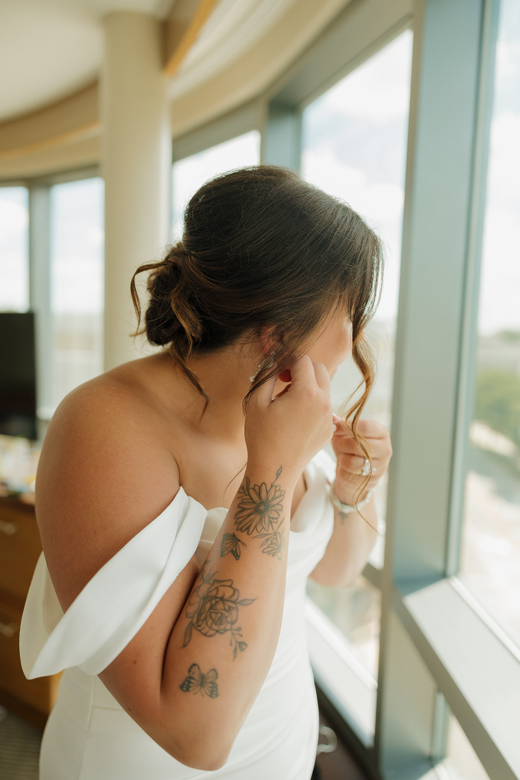 Bride adjusts her earrings near a sunny window, showing off her floral tattoos and soft bridal glam before her Wisconsin brewery wedding.
