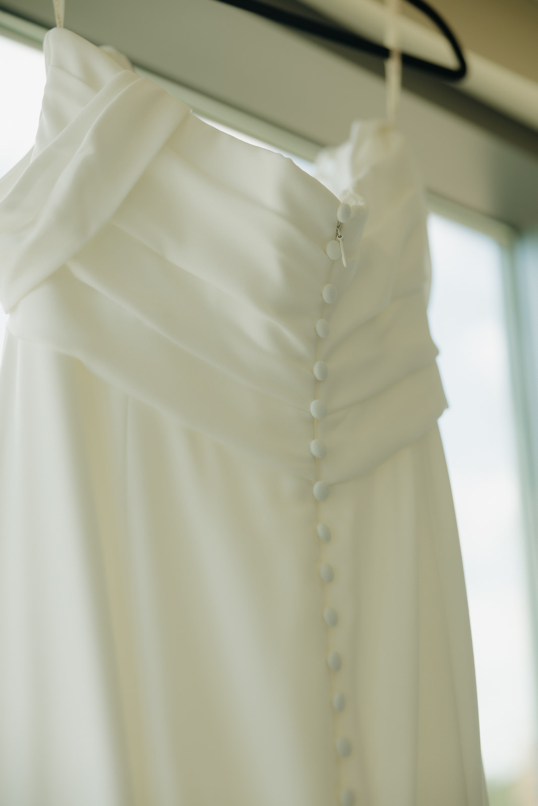 Close-up of an elegant white wedding gown with button detailing, hanging by a window at a Wisconsin brewery wedding venue.