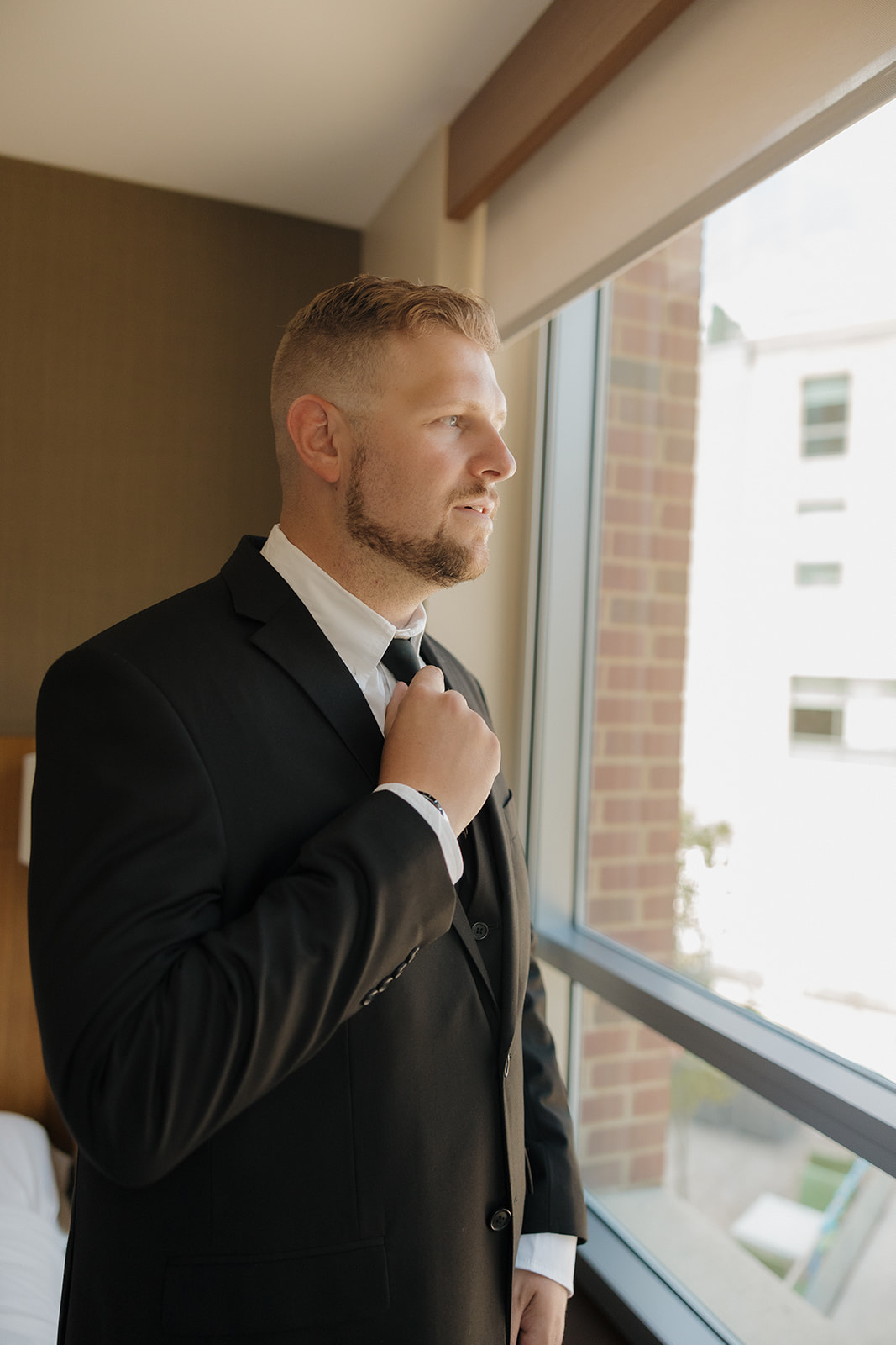 Groom stands near the window adjusting his tie, soaking in a calm moment before his Wisconsin brewery wedding day begins.