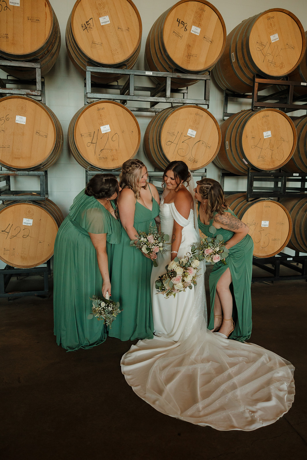 Bride laughs with her bridesmaids in sage green dresses, surrounded by stacked barrels inside a Wisconsin brewery venue.