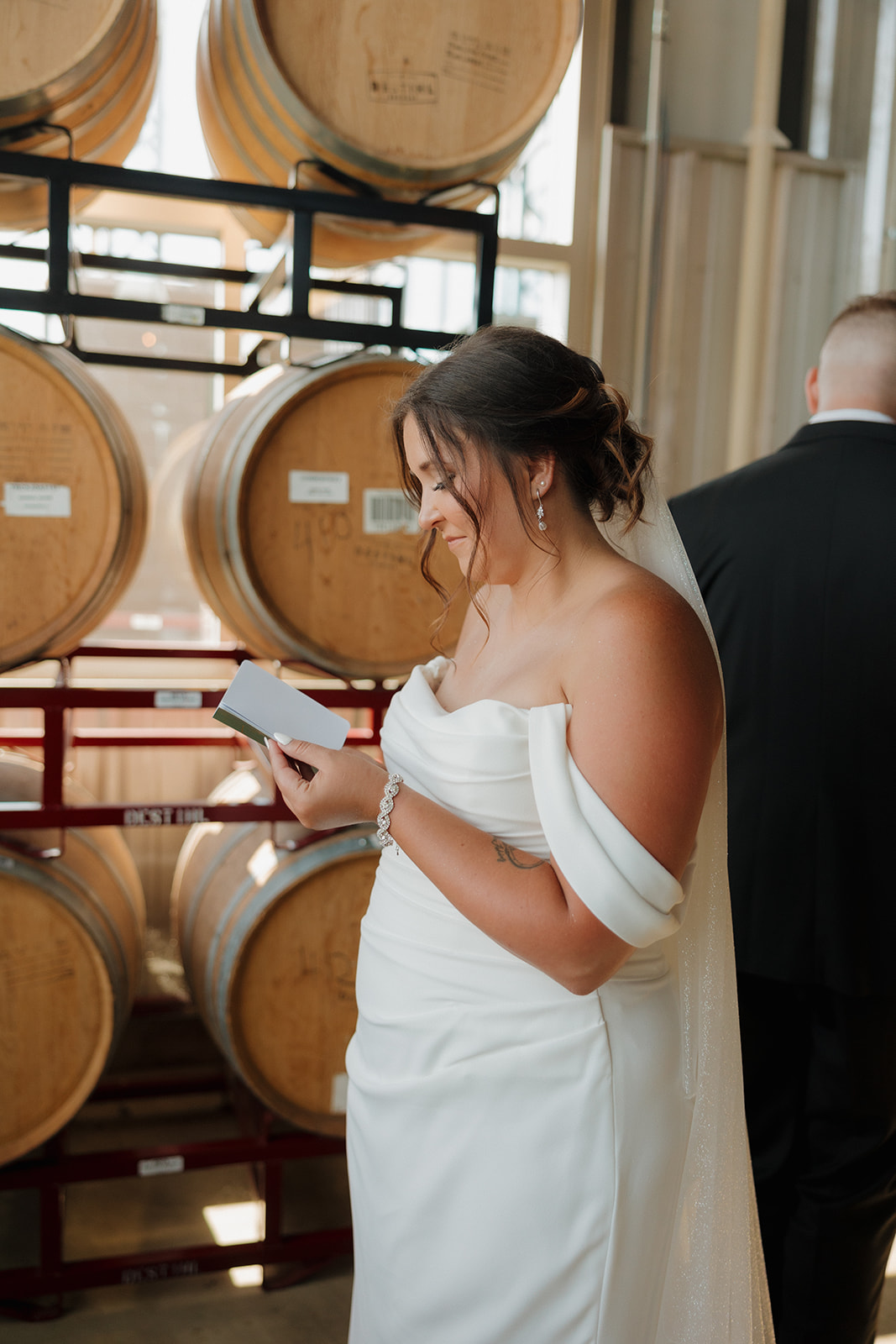 Bride reading her groom’s heartfelt letter with a joyful smile, standing in front of stacked barrels at a Wisconsin brewery.