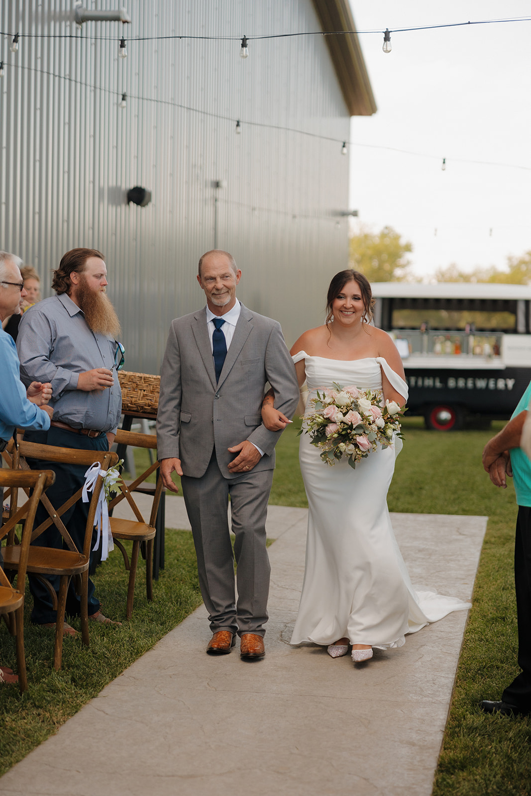 Bride walks arm-in-arm with her father down the outdoor aisle at a Wisconsin brewery wedding, bouquet in hand and glowing.