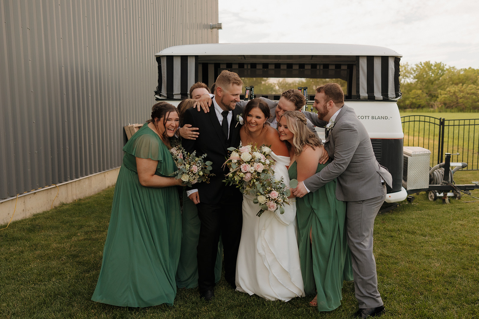 Bride and groom share a group hug with their wedding party in front of a vintage mobile bar setup at a Wisconsin brewery wedding.