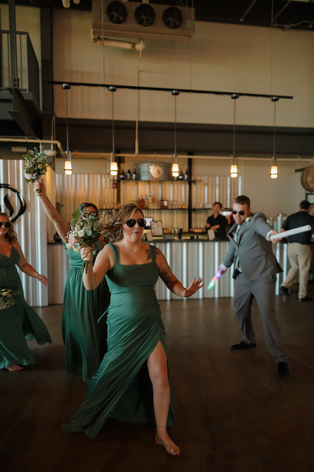 Bridesmaid makes an energetic reception entrance, barefoot and bouquet in hand, dancing through the brewery space.