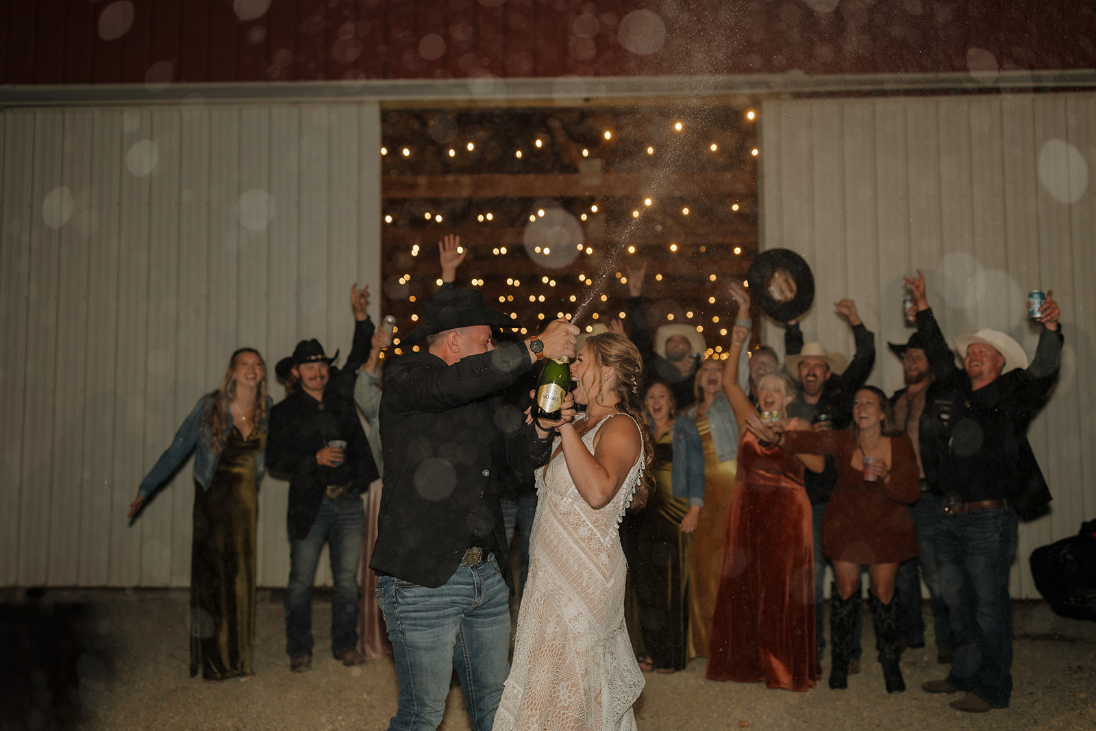 Bride and groom pop champagne and spray it all over while wedding party celebrates behind them