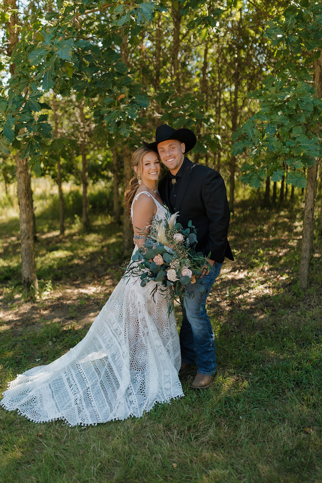Backyard wedding portrait of a smiling bride and groom standing in a shady grove of trees, the bride holding a lush bouquet and the groom in a black cowboy hat.