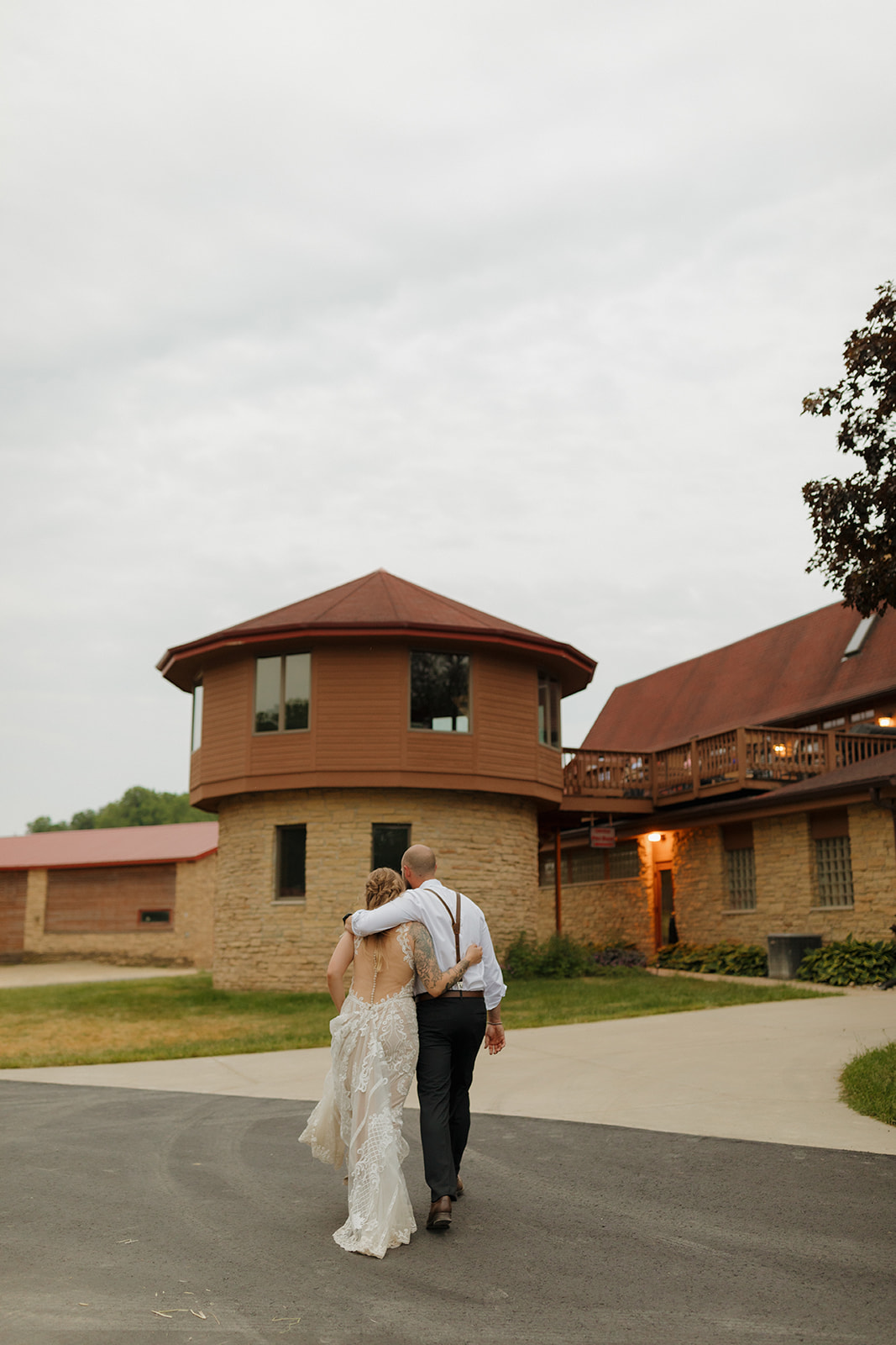 Newlyweds walk away arm-in-arm, their backs to the camera, with a rustic lodge venue in the background and soft clouds above.