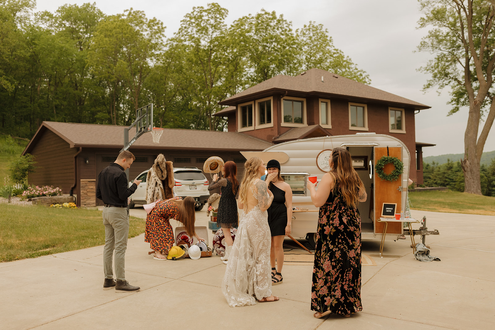Guests gather around a vintage cocktail camper in the driveway of a home, adding charm and casual flair to this backyard wedding celebration.