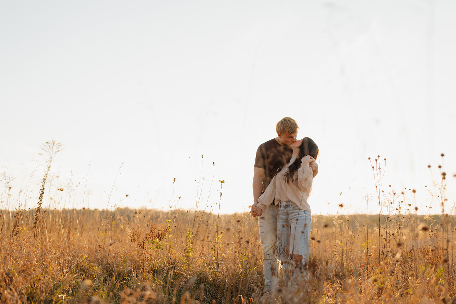 Woman turns her head back to kiss fiance while standing amidst tall golden grass captured by madison wi wedding photographer