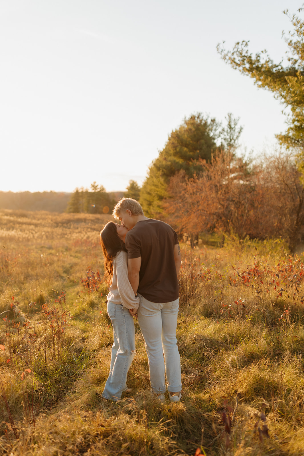 Man leans over to kiss fiance during outdoor engagement photos while holding her hand and walking through field of golden grass
