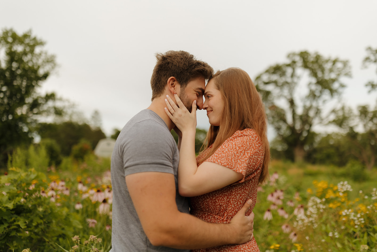 Engaged couple smiling together in a vibrant field of colorful wildflowers under a clear blue sky.