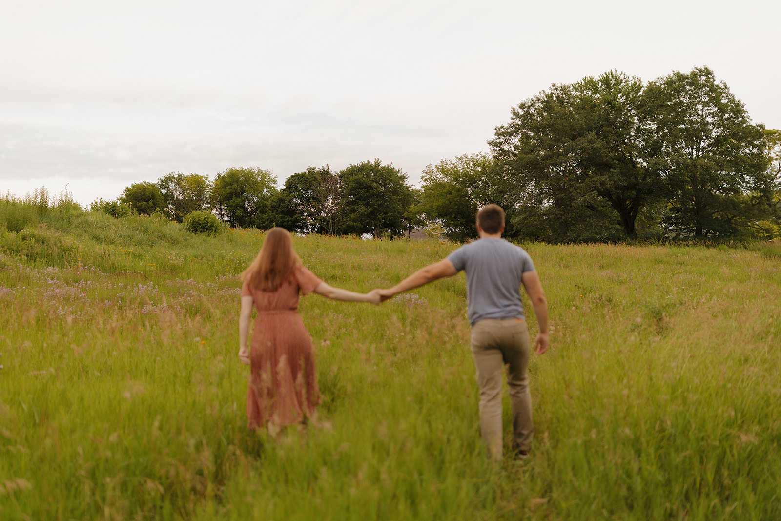 Newly engaged couple holds hands while walking through a field of tall grass at sunset