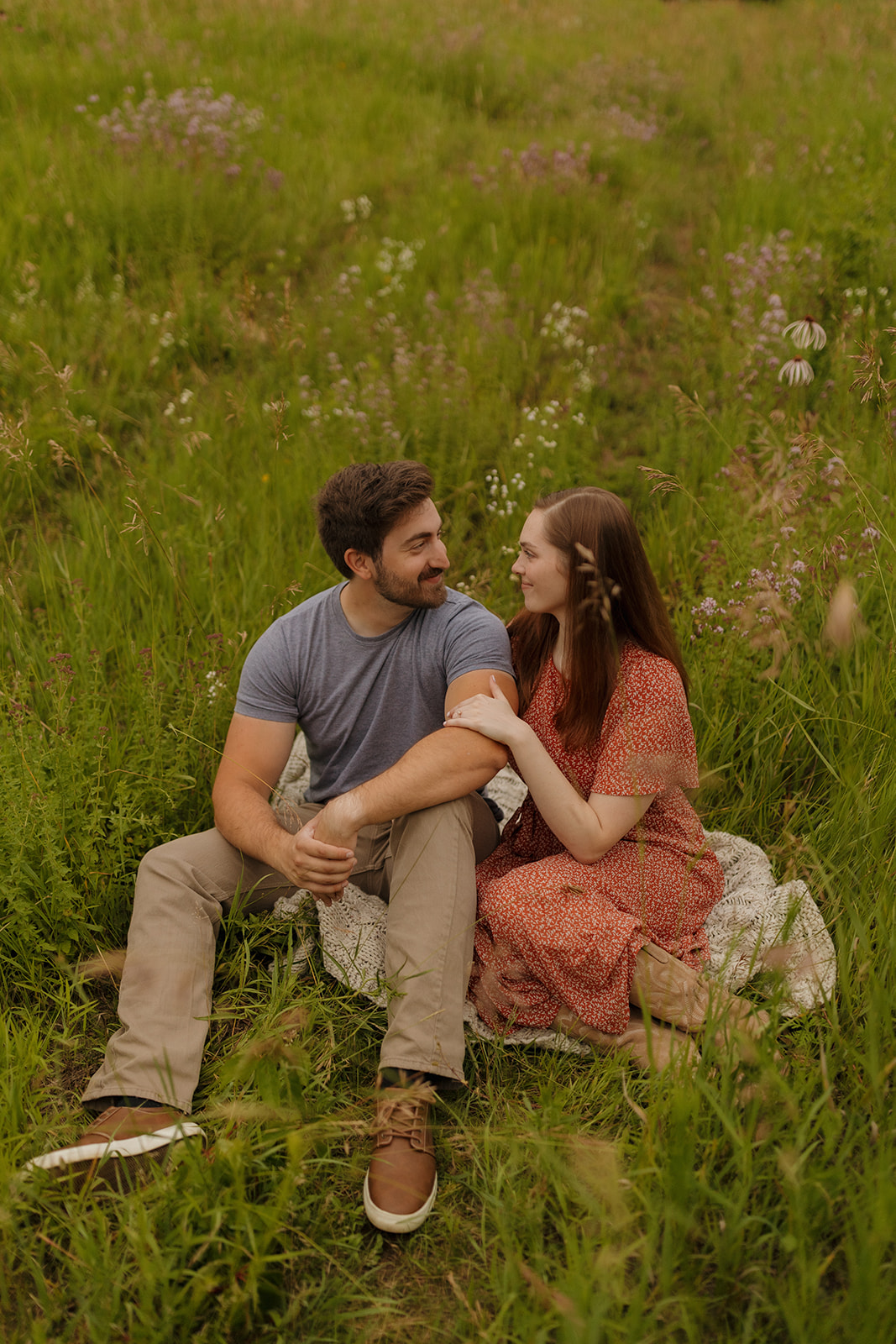 A couple sitting close together on a blanket in a wildflower field, smiling softly at each other on a warm summer evening.