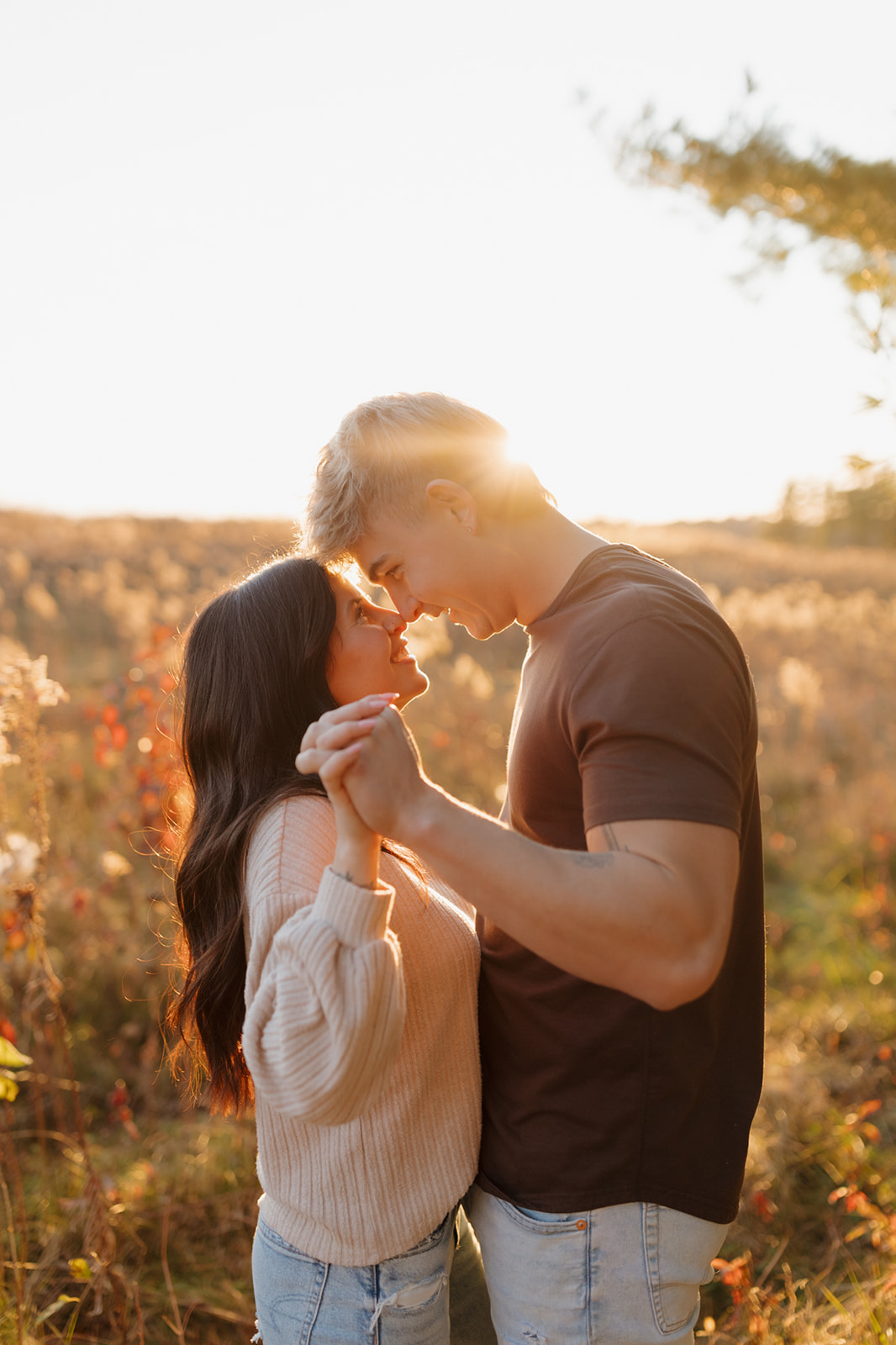 Engaged couple holding hands at sunset during outdoor engagement photos pressing foreheads together and laughing