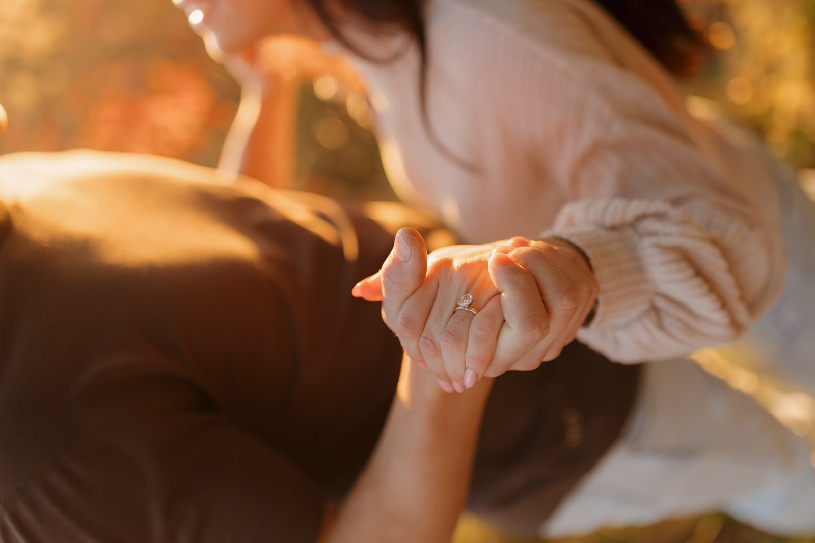 A man and woman clasping hands, basking in the sunlight showcasing woman's engagement ring