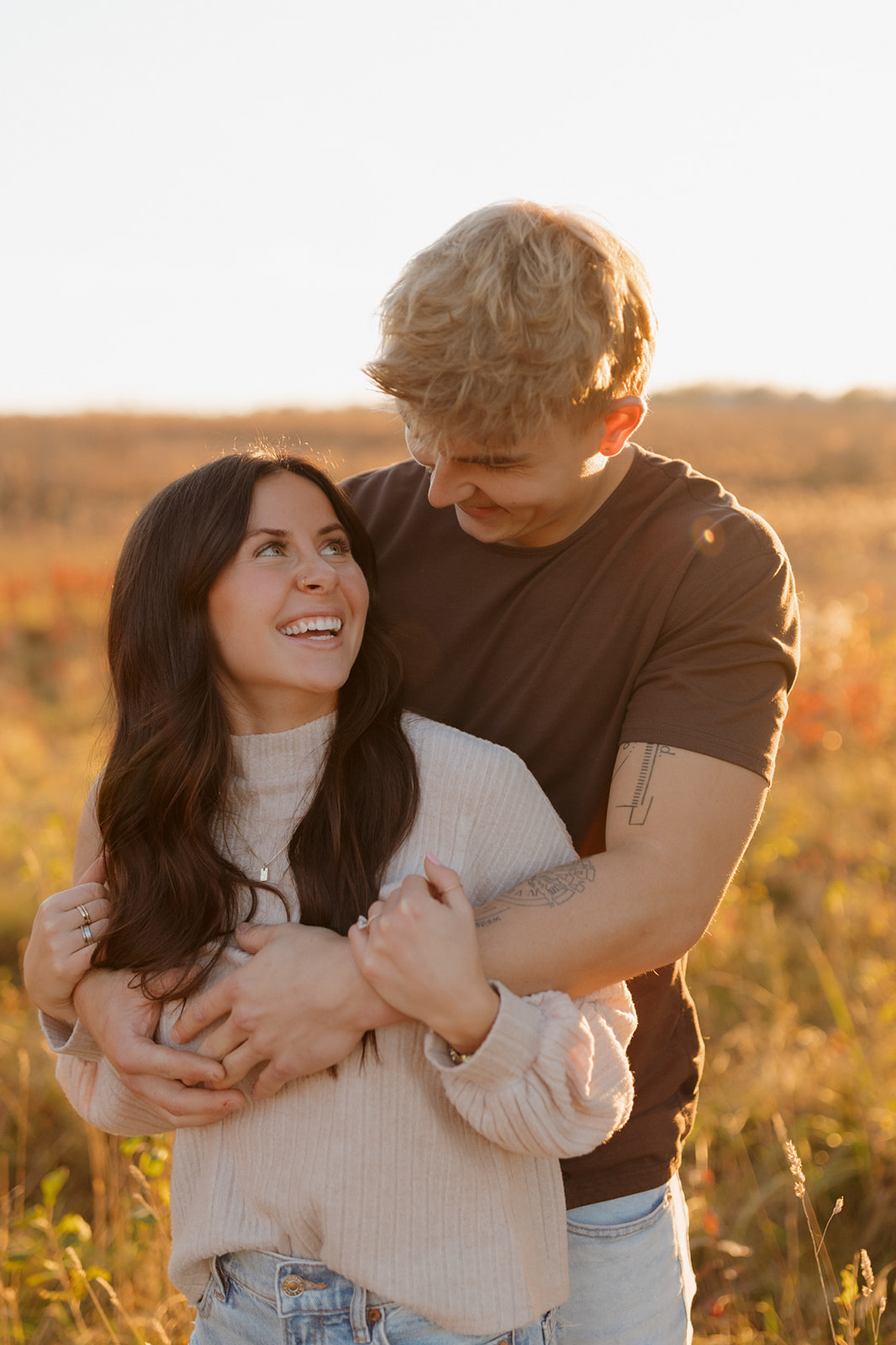 Man holds woman from behind while she looks back laughing at him during golden hour outdoor engagement photos