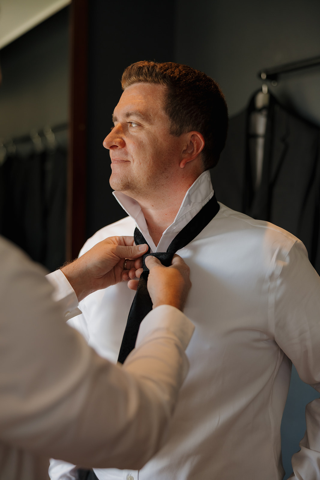 Groom looking away as groomsmen helps him tie his tie before wedding ceremony begins