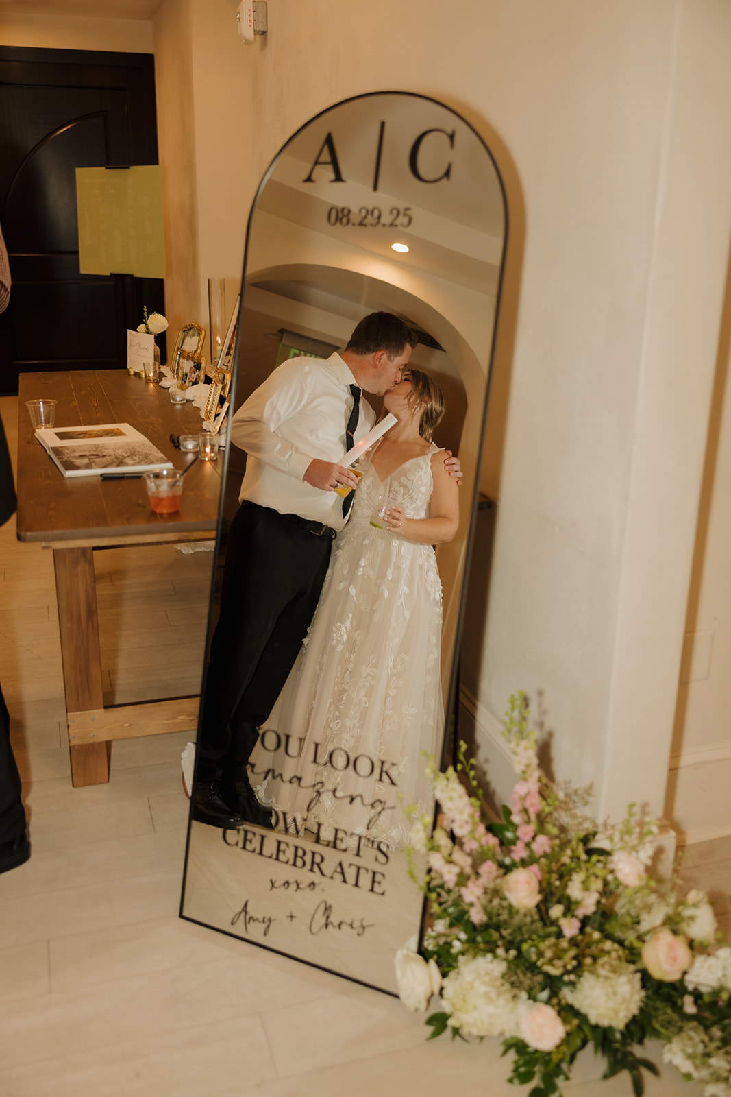 Bride and groom kiss in front of custom mirror with their wedding date holding large glow stick celebrating their marriage