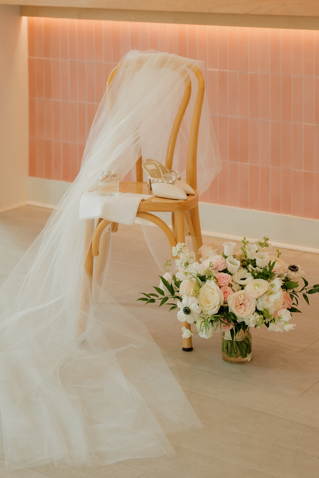 Bridal shoes and veil sitting on wooden chair with bridal bouquet in vase of water before wedding