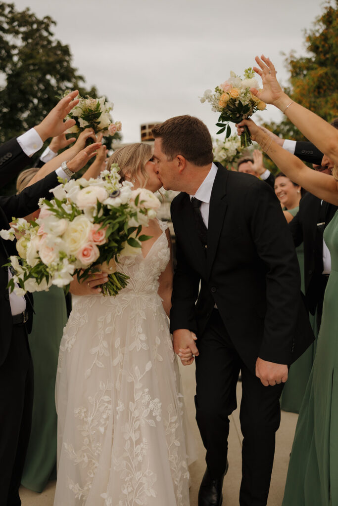 Bride and groom kiss under wedding party's hands while bride holds bouquet after wedding ceremony