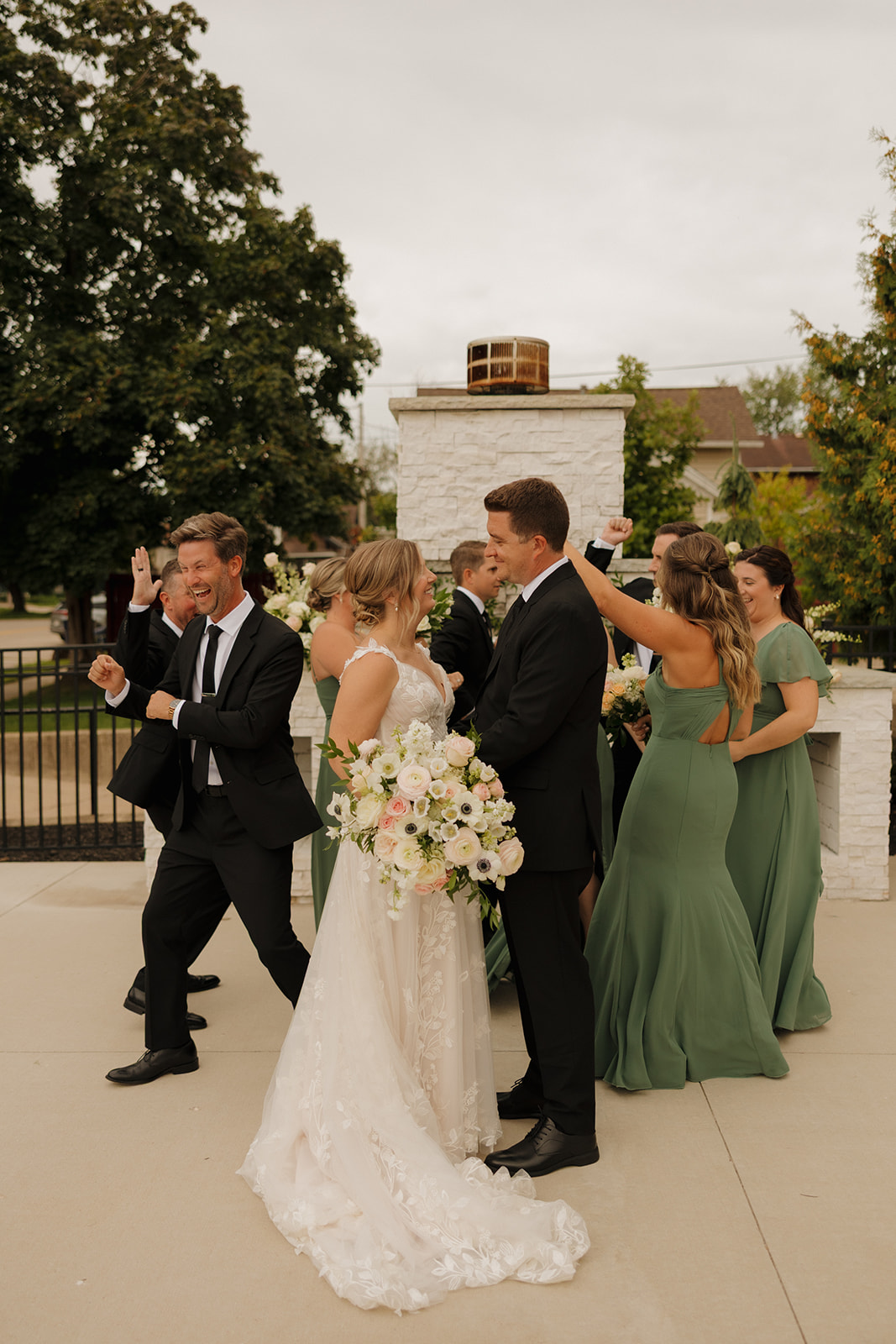 Wedding party dances behind bride and groom celebrating their recent marriage while bride and groom look at one another laughing