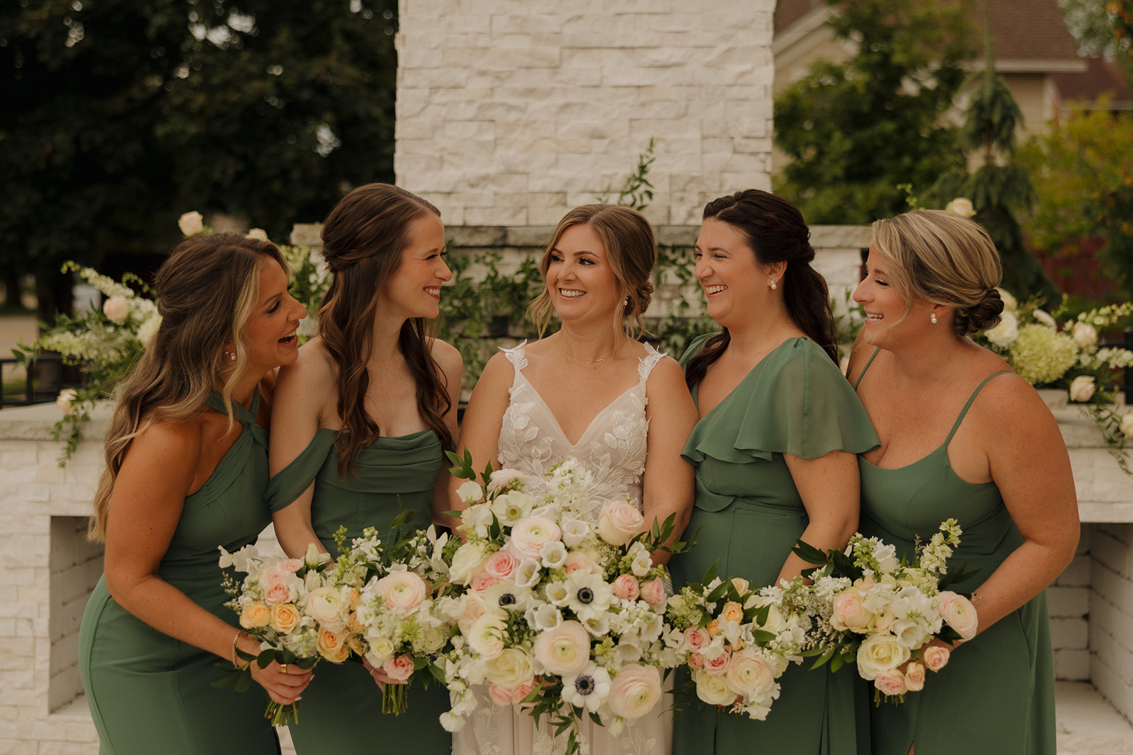 Bride laughing with bridesmaids wearing sage green bridesmaid dresses while holding white and pink bouquets at 10 South wedding venue