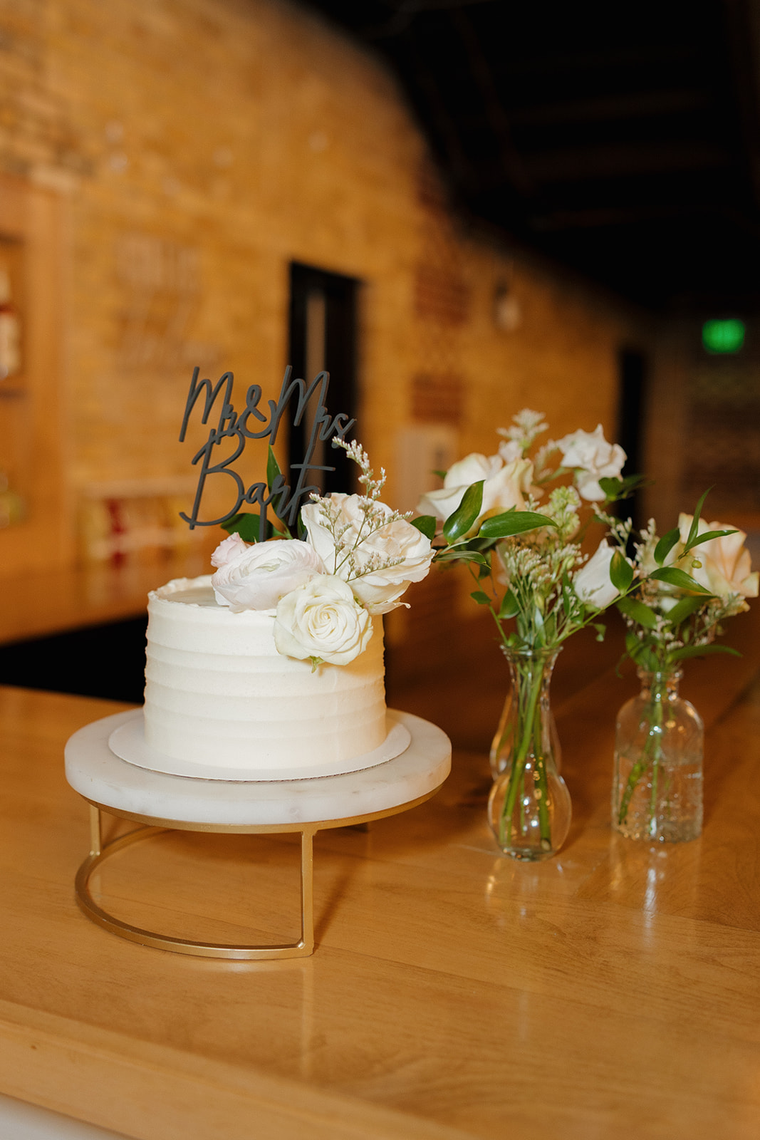 Close up of simple wedding cake with mr and mrs topper sitting beside a simple vase of white flowers and greenery at 10 South wedding venue
