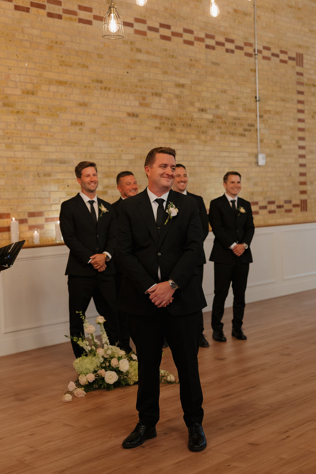 Groom standing at end of aisle looking on as his bride walks down the aisle towards him with a smile on his face. 
